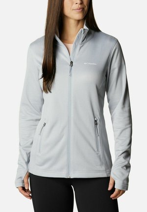 Frau trägt eine hellgraue Columbia-Jacke mit Reißverschluss, Seitentaschen mit Reißverschluss und Daumenlöchern, kombiniert mit schwarzen Hosen.