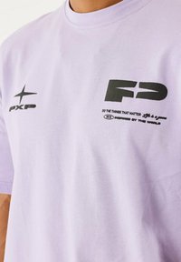 Lichtpaarse katoenen t-shirt met zwarte bedrukte logo's en tekst op de voorkant. Klassieke ronde halslijn en korte mouwen.