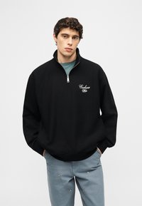 Νεαρός άνδρας με σγουρά μαλλιά, φοράει μαύρο φούτερ με φερμουάρ στο ένα τέταρτο και λευκό λογότυπο, καθώς και μπλε παντελόνι, με τα χέρια στις τσέπες, ουδέτερο φόντο.