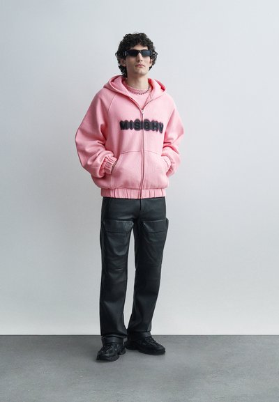 Homme portant des lunettes de soleil noires, un sweat à capuche rose oversize avec le texte "MISBHV", un pantalon cargo noir et des baskets noires, debout contre un mur uni.