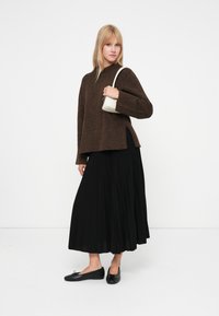 Suéter tricotado castanho texturizado com um decote alto, combinado com uma saia preta plissada e sapatilhas pretas. A segurar uma pequena bolsa branca.