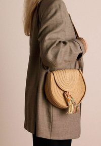 Borsa rotonda in paglia intrecciata con fondo piatto, dotata di chiusura a pattina, frange e accenti decorativi colorati. Tracolla attaccata per il trasporto a spalla.