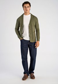 Olivengroen vest met geribbelde details en knoopsluiting, gedragen over een wit overhemd, gecombineerd met donkerblauwe jeans en bruine bootschoenen.