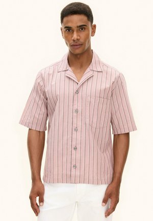 Mann trägt ein kurzärmliges, rosa Hemd mit weißen und schwarzen vertikalen Streifen und dazu eine weiße Hose, steht vor einem einfarbigen Hintergrund.