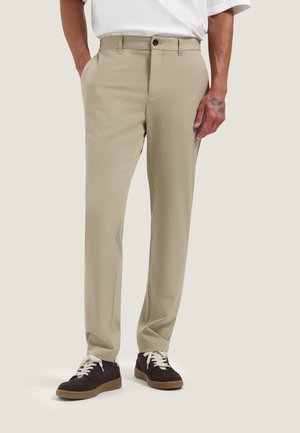 Man wearing beige straight-leg pants with side pockets and black sneakers with white laces, standing against a plain background.