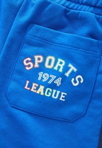 Modré teplákové nohavice majú zadné vrecko ozdobené viacfarebným vyšitým textom: "SPORTS 1974 LEAGUE." Mäkká textúra látky, neformálny dizajn.