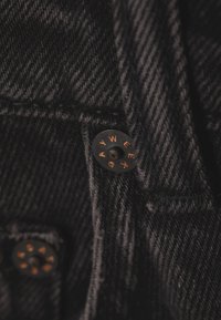 Zwarte denimstof met een gestructureerd patroon. Metalen knoop met reliëfletters: "W E A R", wat duidt op merk of stijl.
