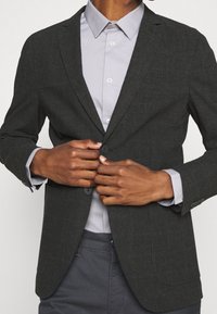 Uhlově šedý blazer s jemným kostkovaným vzorem, se dvěma předními knoflíky a klopami, přeložený přes světle šedou košili.