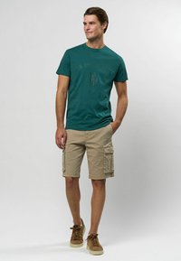 Tīri zaļš kokvilnas T-krekls ar uzrakstu "U.S. Polo Assn.", apvienots ar bēšiem cargo šortiem ar sānu kabatām un brūniem šņorējamiem kedām.