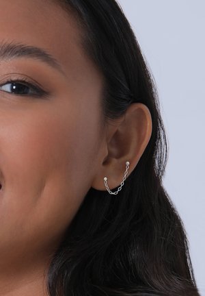 Boucle d'oreille en chaîne argentée avec de petites perles décoratives, présentant un design lié. Portée sur le lobe, elle met en valeur les contours de l'oreille.