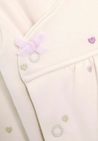 Vêtement pour bébé crème clair avec de petits coeurs brodés en vert, lavande et violet. Il présente un noeud lavande et des détails de fermeture à pression.