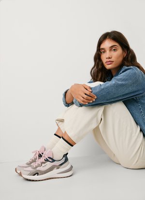 Baskets avec un schéma de couleurs rose et gris, présentant une tige texturée, une semelle blanche et des lacets contrastés. Portées avec un pantalon crème et des chaussettes rayées.