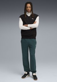 Sort ærmeløs sweater over en hvid turtleneck, grøn sweatpants og sorte sneaks. Har Puma-logo på sweateren og skoene.