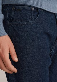 Jins negri din denim cu o finisare texturată, având o buzunar frontal, butoane din metal și o imagine de aproape a unei mâini odihnindu-se pe țesătură.