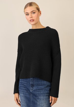 Schwarzer gestrickter Pullover mit geripptem Muster, Bootsausschnitt und langen Ärmeln. Kombiniert mit einem blauen Jeansrock mit Nieten.
