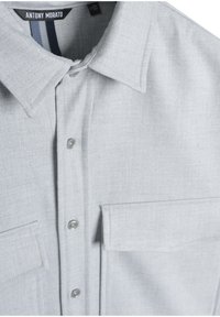 Lichtgrijze button-up shirt gemaakt van textuurstof. Bevat een puntige kraag, twee borstzakken en doorzichtige plastic knopen.