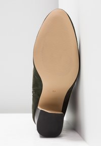 Semelle de chaussure en daim vert avec dessous beige texturé et talon bloc noir, taille 37, inclinée contre un fond blanc d’angle.