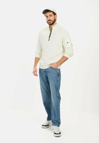 Weißer Reißverschluss-Pullover mit gerippter Textur, kombiniert mit hellblauen Jeans. Eine leichte Mütze mit kontrastierendem Schirm rundet das Outfit ab.