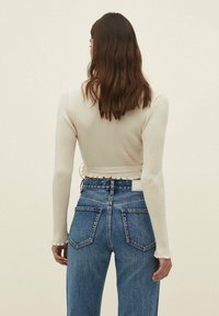 Valkoinen ribattu pitkähihaine crop-top, jossa on röyhelöhelma ja löysät hihat, yhdistettynä korkeavyötäröisiin sinisiin farkkuihin, joissa on takataskut.