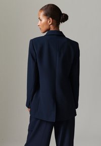 Granatowa marynarka o dopasowanym kroju, z klapami z wycięciem i usztywnionym tyłem. Materiał wydaje się gładki o matowym wykończeniu.