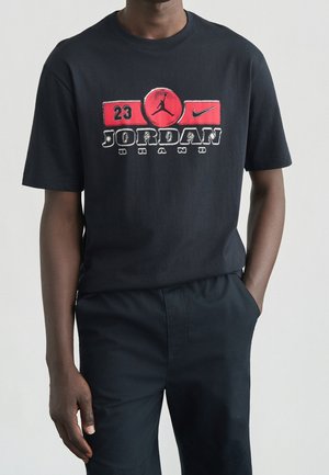 Person iført sort Jordan Brand t-shirt med røde og hvide logoer, nummer 23, og Nike swoosh, klædt i sorte bukser.