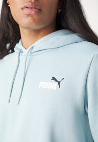 Vaaleansininen huppari, jossa on kengurutasku, nyörikiinnitteinen huppu ja musta Puman logo rinnassa. Pehmeä kangas, jossa on sileä pinta.