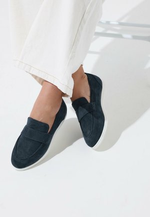 Persona con mocasines negros de ante sin cordones con suelas blancas, combinados con pantalones blancos cropped, sentada sobre una superficie blanca.