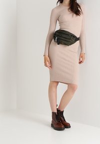 Robe en maille côtelée beige à manches longues, silhouette ajustée, associée à une banane vert olive et des bottes de combat marron à lacets.