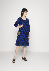 Vestido azul com estampado, decote quadrado e mangas três quartos. Combinado com mules pretos e uma pequena bolsa de ombro.