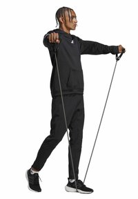 Schwarzer Hoodie mit einer Fronttasche, kombiniert mit schwarzen Jogginghosen und schwarzen Sneakers. Der Benutzer hält zwei Sportstöcke und steht in einer dynamischen Pose.