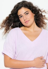 Top viola chiaro con scollo a V e maniche arrotolate, realizzato in tessuto texturizzato. Modella con capelli ricci e orecchini a cerchio, che tiene il braccio.
