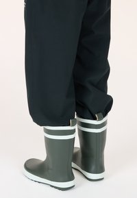 Bottes de pluie en caoutchouc vertes avec des rayures blanches, dotées d'une semelle robuste et d'une languette à l'arrière. Portées avec un pantalon étanche noir.