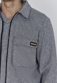 Cămașă gri cu model în herringbone, dotată cu fermoar, având un buzunar pe piept și o etichetă mică cu brand. Țesătură texturată și guler cu nasturi.