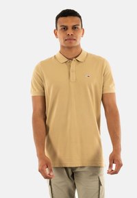 Polo beige con cuello clásico, manga corta y un pequeño logo en el pecho. La tela parece suave y ligera.