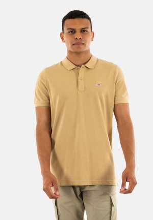 Polo beige avec un col classique, des manches courtes et un petit logo sur la poitrine. Le tissu semble lisse et léger.