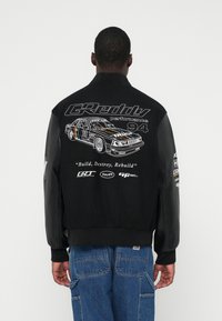 Fekete bomberdzseki bőr ujjakkal, versenygrafikával és szöveggel a hátoldalon, benne az "Építs, pusztíts, építs újjá" felirattal.