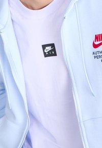 Sudadera con cremallera azul claro sobre una camiseta lila, que presenta una etiqueta cuadrada negra con "NIKE AIR" y acentos de logotipo en rojo en la sudadera.