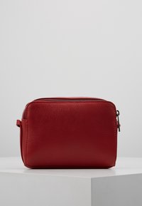 Punainen nahkalaukku, suorakulmainen muoto, teksturoitu pinta ja yläosan vetoketjusuljenta. Varustettu pienellä sivukahvalla helppoa kantamista varten.