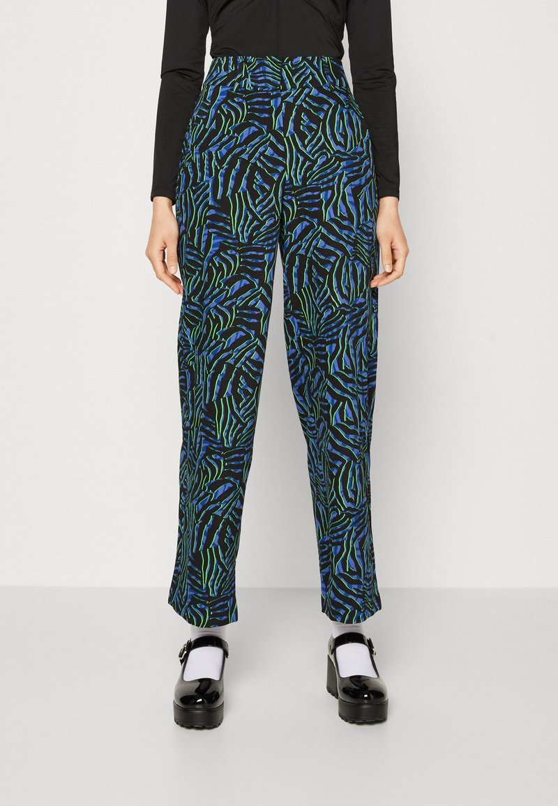 Femme portant un pantalon taille haute avec des rayures zèbre abstraites bleues, vertes et noires et des chaussures Mary Jane noires avec des chaussettes blanches.