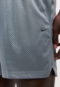 Gros plan sur un short de sport gris perforé avec un petit logo Nike noir, porté par une personne à la peau foncée tenant le côté du short.