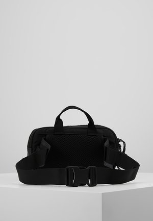 Sort kompakt crossbody taske med netpanel på bagsiden, justerbar bred rem og top-håndtag, vist på en hvid overflade.