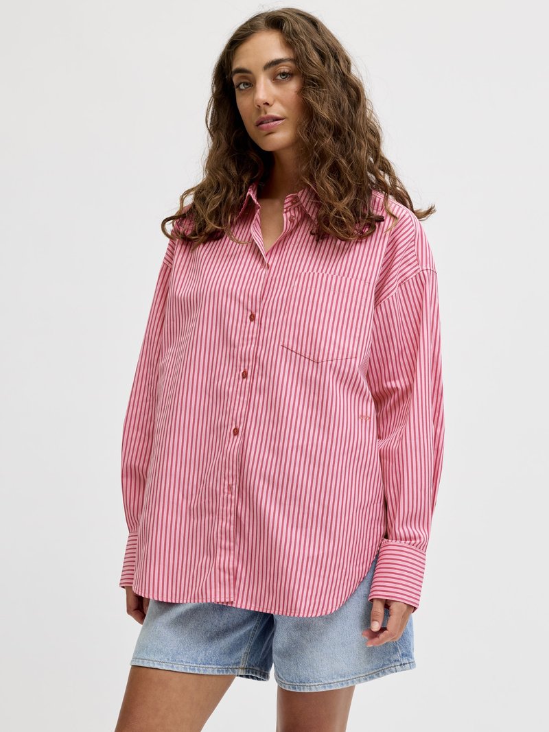 Donna con lunghi capelli ricci che indossa una camicia oversize a righe rosa e bianche con bottoni e pantaloncini di jeans azzurri chiari.