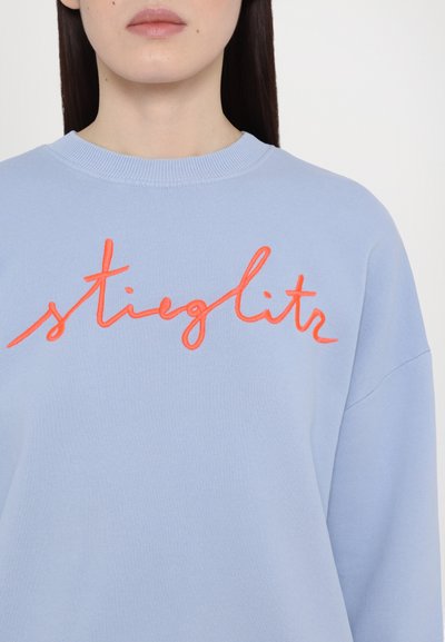 Light blue sweatshirt featuring orange embroidered script reading "stieglitz." The fabric appears soft and textured with a classic crew neckline.