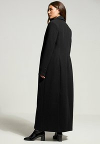 Cappotto lungo nero realizzato in lana texture con silhouette su misura, colletto a scollatura e polsini abbottonati, che arriva a metà polpaccio.