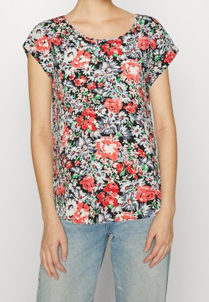 Kobieta ubrana w bluzkę z krótkim rękawem w czerwony, szary i biały kwiatowy wzór, zestawioną z jasnoniebieskimi dżinsami.