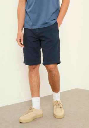 Man wearing navy blue shorts, a blue shirt, white socks, and beige sneakers standing indoors on a gray floor.
