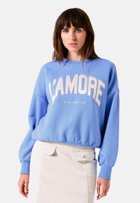 Sudadera azul claro con "L’Amore" en letras grandes y en contraste. Presenta puños y dobladillo acanalados. Combinada con una falda de pana beige.