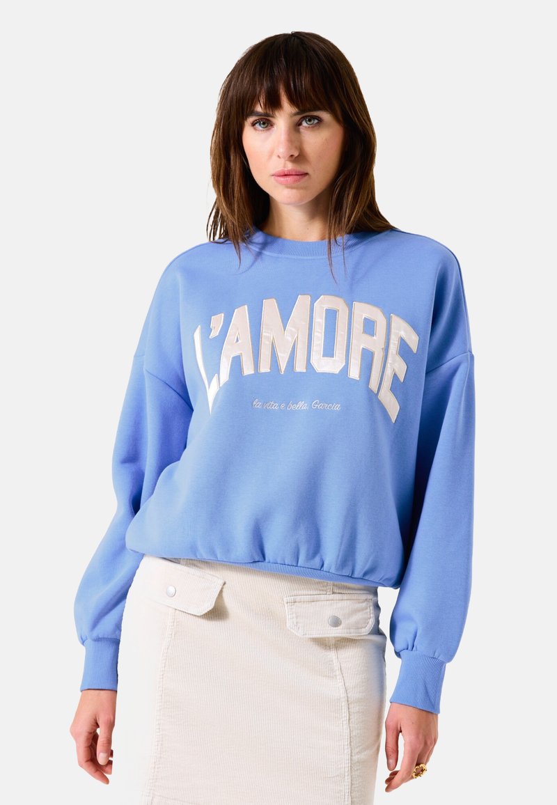 Sudadera azul claro con "L’Amore" en letras grandes y en contraste. Presenta puños y dobladillo acanalados. Combinada con una falda de pana beige.