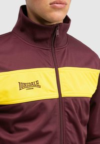Burgundi színű, cipzáros dzseki magas gallérral, vízszintes sárga panelekkel és barna "Lonsdale London" logóval a mellkason.