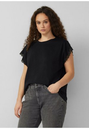 Frau mit lockigem Haar, die ein schwarzes T-Shirt mit spitzenbesetzten Ärmeln und graue Jeans trägt, steht vor einem einfarbigen grauen Hintergrund.